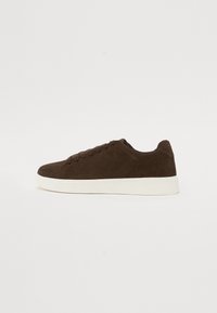 Brown suede sneaker with a low profile, featuring a white rubber sole, round toe, and six eyelets for laces. Smooth texture and simple design.