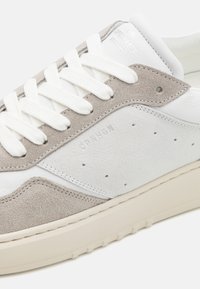 Sneaker featuring a combination of white leather and gray suede, flat white laces, and a cushioned beige sole. Subtle perforated detailing.