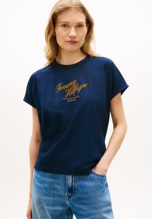 Νεαρή γυναίκα με ανοιχτόχρωμα κυματιστά μαλλιά φοράει μπλε σκούρο T-shirt Tommy Hilfiger και ανοιχτόχρωμο τζιν, με το ένα χέρι στην τσέπη.