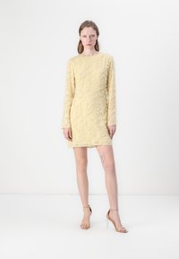 Gelbes Mini-Kleid mit langen Ärmeln, strukturiertem Stoff und schichtigen Fransen-Details. Das Model trägt beigefarbene Ankle-Strap-High Heels.