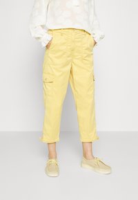 Vrouw in een korte gele cargobroek met voor- en zijzakken, gecombineerd met beige suède schoenen en een witte blouse met een bloemmotief.