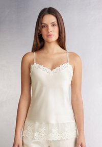 Satin camisole in licht ivoor met verstelbare bandjes, kantafwerking bij de halslijn en zoom, met een gladde textuur en een delicate bloemenpatroon.