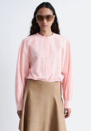 Kobieta w dużych brązowych okularach przeciwsłonecznych, jasnoróżowej koszuli zapinanej na guziki i beżowej spódnicy o kroju litery A, stojąca na jednolitym tle.