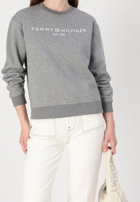 Szara bluza z okrągłym dekoltem i prążkowanymi mankietami, z białym napisem "TOMMY HILFIGER EST. 1985" na przodzie. Białe spodnie i torba.