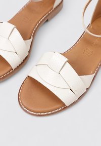 Sandales en cuir blanc avec un design de bride tressée, semelle plate couleur tan et détails brodés. Texture lisse et style à bout ouvert.