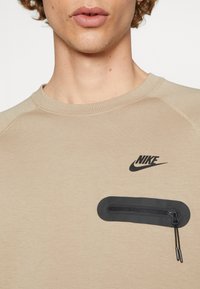 Beżowa bluza Nike z czarnym logo i poziomą kieszenią na zamek błyskawiczny na klatce piersiowej, noszona przez osobę z kręconymi włosami.