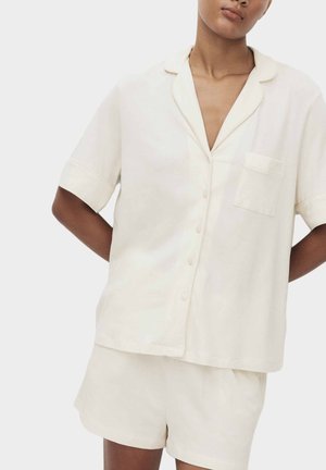 Model draagt een losvallend, kort mouwen crèmekleurig overhemd met kraag en zak, gecombineerd met bijpassende crèmekleurige shorts tegen een effen achtergrond.