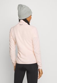 Světle růžový dlouhý rukáv top s vysokým límcem, vyrobený z měkké látky. Obsahuje malé logo na zadní straně a přiléhavý střih. Kombinováno s černými legínami.