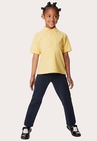 Žluté polotriko s krátkými rukávy, doplněné tmavě modrými kalhotami a černými botami. Košile má límeček a zapínání na knoflíky.