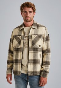 Ruitenfleecejack met beige en bruine kleuren, twee borstzakken en zwart patchdetail op de linkerarm. Draagt over een licht T-shirt.