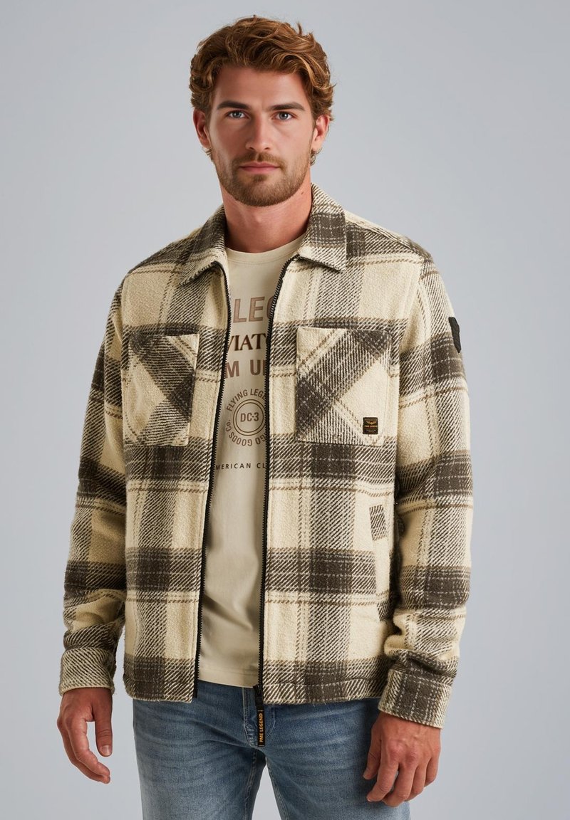 Ruitenfleecejack met beige en bruine kleuren, twee borstzakken en zwart patchdetail op de linkerarm. Draagt over een licht T-shirt.