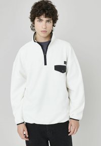 Pull en polaire blanche avec une fermeture �éclair noire, col montant et poche poitrine. Présente des manches amples et une texture douce.