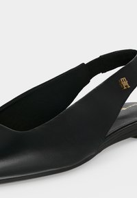 Μαύρο δερμάτινο flat παπούτσι με λουράκι στην φτέρνα, μυτερή μύτη και μικρό χρυσό λογότυπο στο πλάι.