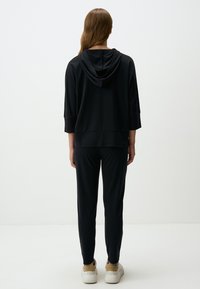 Felpa nera con tasca frontale, maniche a tre quarti e tessuto testurizzato. Indossata con pantaloni neri a sigaretta coordinati e scarpe bianche.