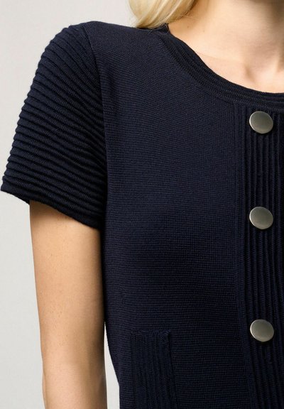 Top lavorato a maglia blu navy con maniche corte a coste, dotato di un collo rotondo e bottoni argentati sul davanti. Include una tasca laterale.