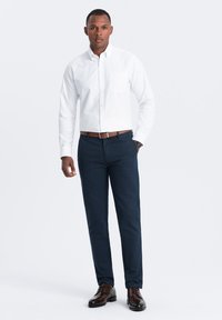 Camicia bianca a maniche lunghe con bottoni, pantaloni blu navy, cintura in pelle marrone e scarpe scure. La camicia ha una tasca sul petto; abbigliamento formale.