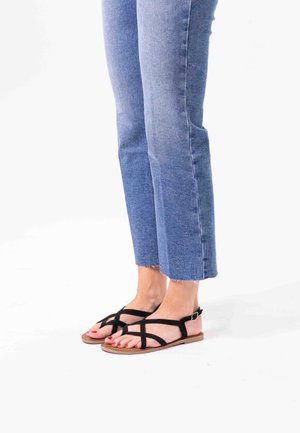 Black suede sandals with crisscross straps, adjustable buckle, and flat tan sole, paired with light blue cropped denim jeans.