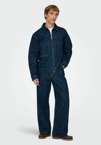 Temno modra denim kombinezon s sprednjim zadrgo, dvema prsna žepa in širokimi hlačnicami. Tkanina ima teksturiran videz in vidne šive.