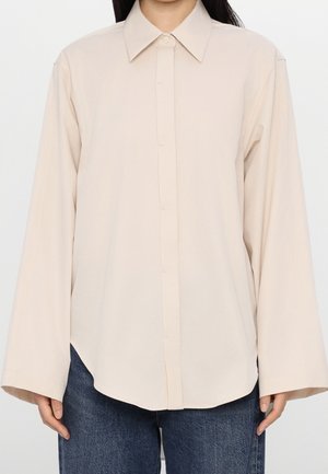 Person iført en løs beige langærmet skjorte med knapper kombineret med blå denimjeans, stående mod en ensfarvet baggrund.