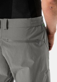 Pantalones exteriores grises con cintura elástica, un bolsillo trasero con cremallera y una textura suave. Con un pequeño etiqueta de logo verde como acento.