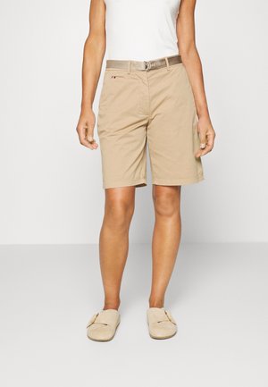 Person iført beige knælange shorts med matchende bælte, hvid ærmeløs top og beige slip-on sko, stående mod en ensfarvet baggrund.