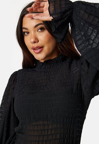 Blusa nera textured con un alto collo arricciato e maniche lunghe a campana, con un aspetto trasparente e un design di tessuto arricciato.