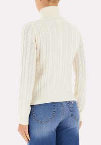 Maglione a treccia color crema con collo alto e orlo a coste, caratterizzato da un motivo testurizzato. Indossato con jeans blu, che mostrano un design attillato.