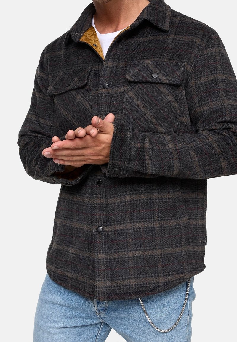Koszula flanelowa w kratę w ciemnoszarym kolorze, z subtelnymi czerwonymi i brązowymi detalami. Posiada guziki na piersi oraz kontrastowe żółte podszewki.