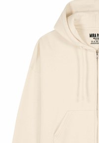 Sweat à capuche crème avec fermeture éclair, poche avant, capuche à cordon de serrage, texture de tissu lisse et design épuré. Étiquette de la marque visible à l'intérieur.