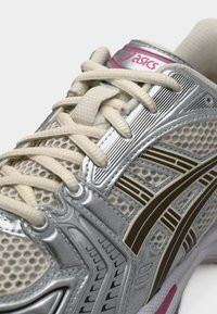 Ασημένιο αθλητικό παπούτσι με δίχτυ στο πάνω μέρος, κρεμ κορδόνια και μαύρες και χρυσές λεπτομέρειες. Διαθέτει υφή στην επιφάνεια και λογότυπο στη γλώσσα του παπουτσιού.