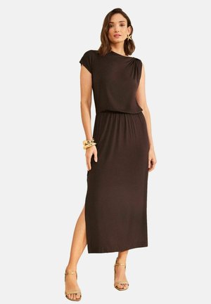 Frau trägt ein ärmelloses schwarzes Midikleid mit seitlichem Schlitz, goldene Creolen, auffällige Armbänder und goldene Sandalen mit hohen Absätzen, stehend.