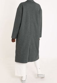 Cappotto grigio a spina di pesce con vestibilità rilassata, maniche lunghe e dettaglio sul retro a spacco, abbinato a pantaloni bianchi e scarpe a righe.
