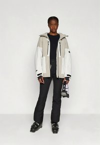 Μπουφάν για σκι από ελαφρύ ύφασμα σε μπεζ και λευκό, με κουκούλα, τσέπες με φερμουάρ και μαύρες λεπτομέρειες. Φοριέται με μαύρο παντελόνι για σκι και μπότες.