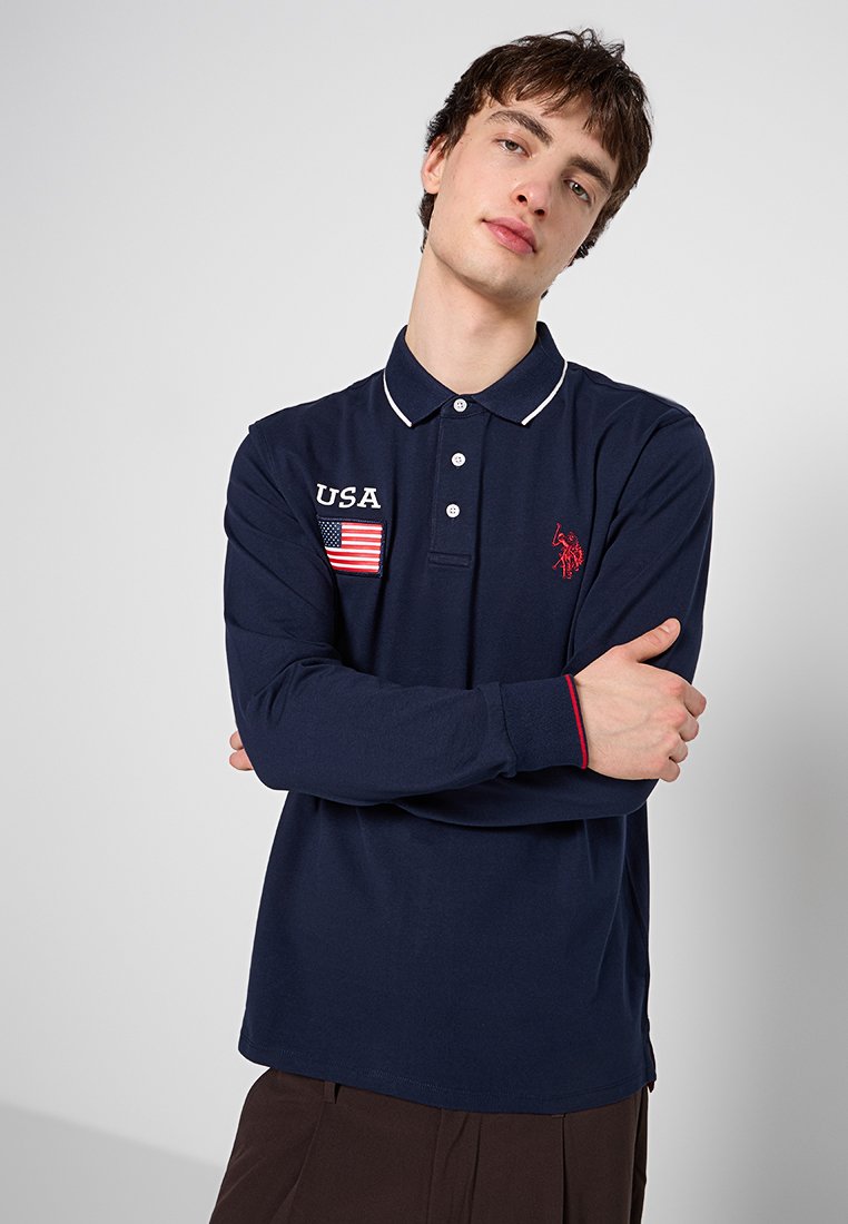 Polo de manga larga en color navy, material de algodón, con ribete blanco en el cuello. Presenta un parche con la bandera de EE. UU. y un logotipo bordado en rojo. Diseño de ajuste clásico.