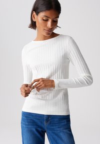 Weißes, geripptes Langarmoberteil mit weitem Ausschnitt, das eine weiche Textur und eine taillierte Passform aufweist, kombiniert mit blauen Jeans in einem lässigen Setting.