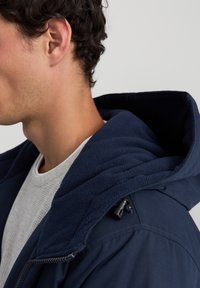 Giacca blu navy con cappuccio foderato in pile, tessuto testurizzato e chiusura a toggle sicura. Il modello indossa una maglietta bianca a maniche corte sotto.