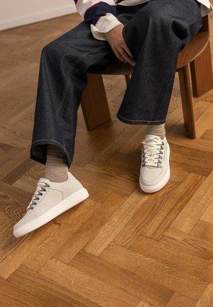 Personne assise sur une chaise en bois portant un jean large foncé, des chaussettes beiges et des baskets blanches à lacets sur un sol en parquet.