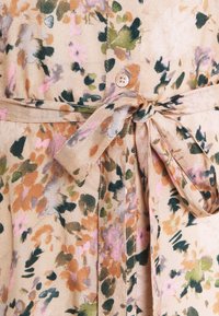 Robe à motif floral beige avec des accents rose, vert et orange. Caractérisée par une ceinture en tissu nouée et une fermeture à boutons au niveau du col.