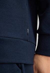 Navy blue sweatshirt with ribbed cuffs and hem, featuring a small black tag with "JOOP!" near the seam, made from a soft, textured fabric.