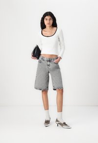 Hvit langermet topp med svart nakke detaljer, kombinert med grå denimshorts og sølvfottøy, mens hun holder en svart håndveske. Enkelt bakgrunn.