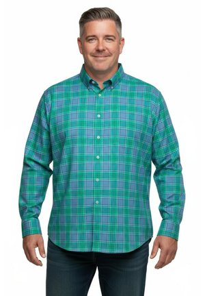 Lächelnder Mann mit einem langärmligen grün-blau karierten Hemd und dunklen Jeans, stehend vor weißem Hintergrund.