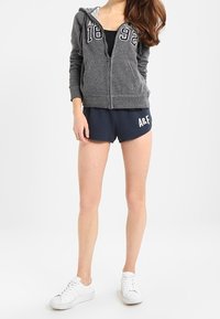 Sudadera gris con cremallera y letras blancas "A&F 1892", combinada con pantalones cortos azul marino que tienen el logo de "A&F". Llevado con zapatillas blancas.