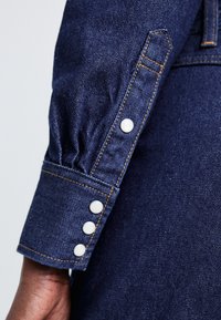 Manșetă de cămașă din denim de culoare albastru închis, cu trei nasturi albaștri și cusături portocalii în contrast. Prezintă material strâns la manșetă.