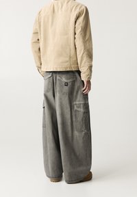 Beige manchesterjacka med rak passform, i kombination med lösa grå cargobyxor med stora sidofickor och en logotyp på baksidan.