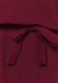 La tela del vestido burdeos presenta una textura suave con un detalle de cinturón atado en la cintura y suaves pliegues para una silueta fluida.
