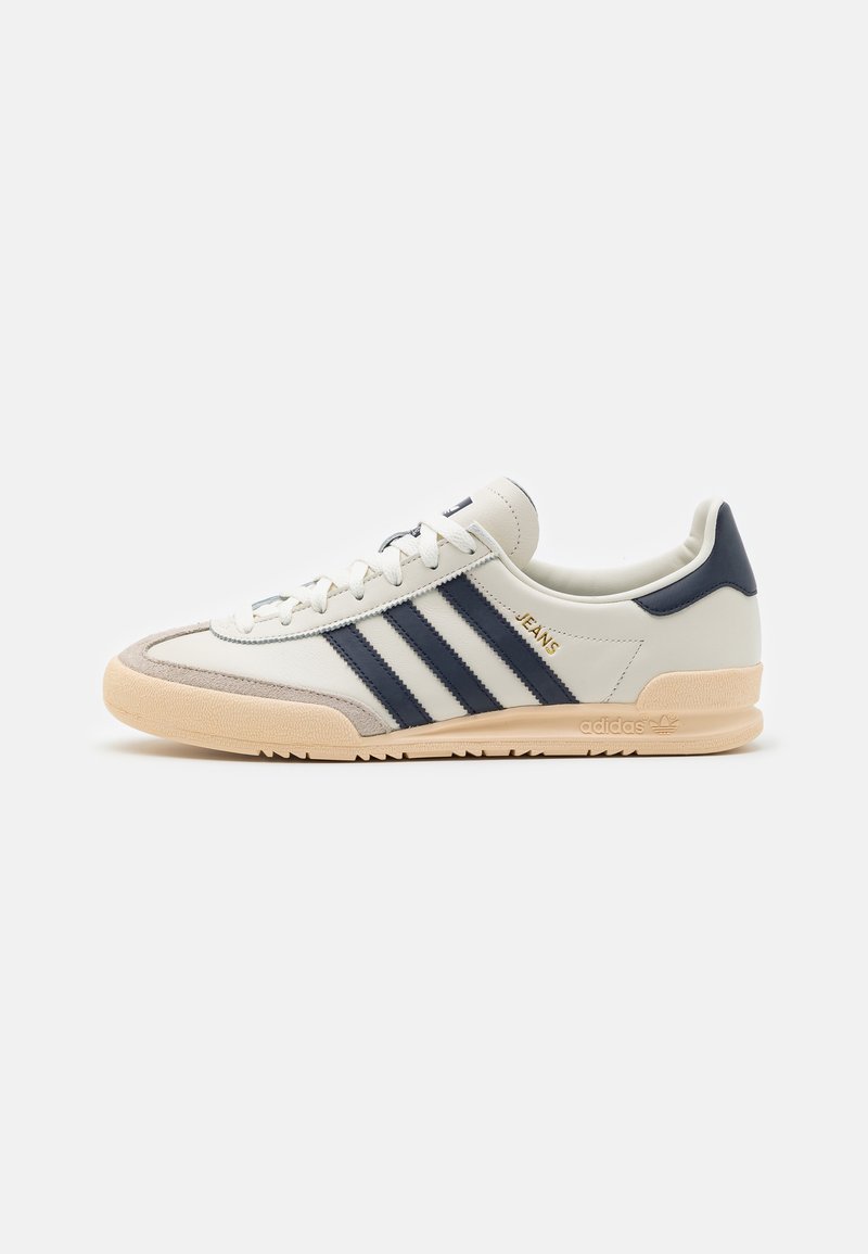 Baskets en cuir blanc avec des rayures bleu marine, accents en daim et semelle en caoutchouc. Elles présentent un design à lacets et un logo doré sur le côté.
