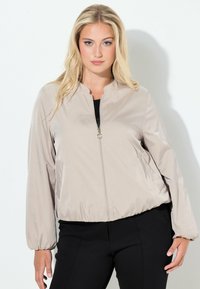 Béžová bomber bunda se zipem vpředu, elastickým pasem a dlouhými rukávy. Hladká látka s mírně lesklým povrchem. Nošena přes černé tričko a kalhoty.
