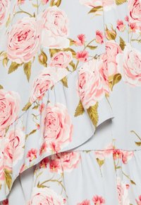 Détail d'une robe fleurie en tissu bleu clair avec des roses roses et des feuilles vertes, ornée de volants aux bords des coutures. Texture lisse.