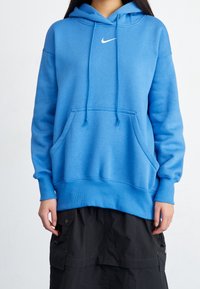 Blå hoodie gjord av mjukt material, med en känguruficka, huva med dragsko och vit Nike-logotyp. Ribbstickade muddar och fåll.