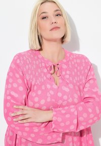 Rosa Bluse mit großen weißen Punkten, einem Schlüsselloch-Ausschnitt mit Bändern und strukturiertem Smocking am Oberteil. Lange, fließende Ärmel.
